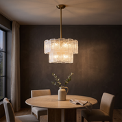Eirene two-tier brass and textured ice-glass chandelier lit over a travertine table in a dark limewash dining room